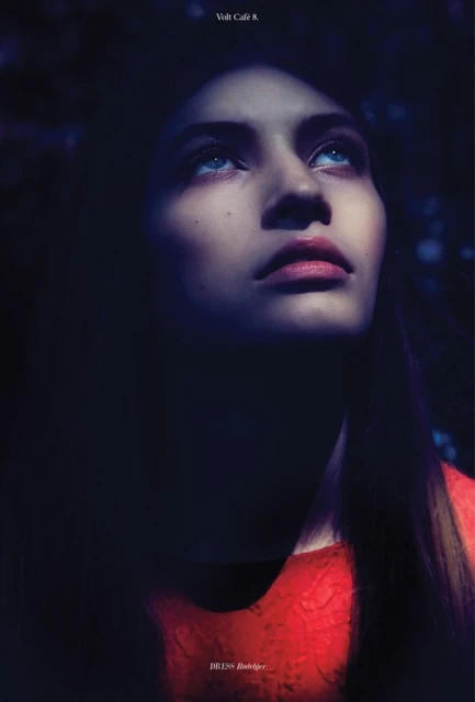 Portrait of a fair-skinned young woman in a red dress looking up with light blue eyes in a dark, shadowy outdoor environment.