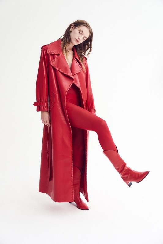 A female fashion model strikes a pose wearing a red monochromatic outfit that consists of a long trench coat, bodysuit, tights, and heeled boots.