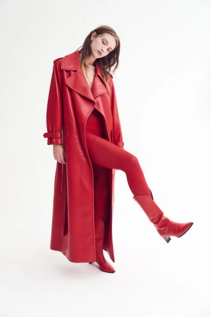 A female fashion model strikes a pose wearing a red monochromatic outfit that consists of a long trench coat, bodysuit, tights, and heeled boots.