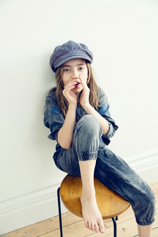 A young girl with long hair, wearing a blue denim jumpsuit and a striped cap, poses indoors while sitting on a wooden stool.