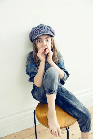 A young girl with long hair, wearing a blue denim jumpsuit and a striped cap, poses indoors while sitting on a wooden stool.