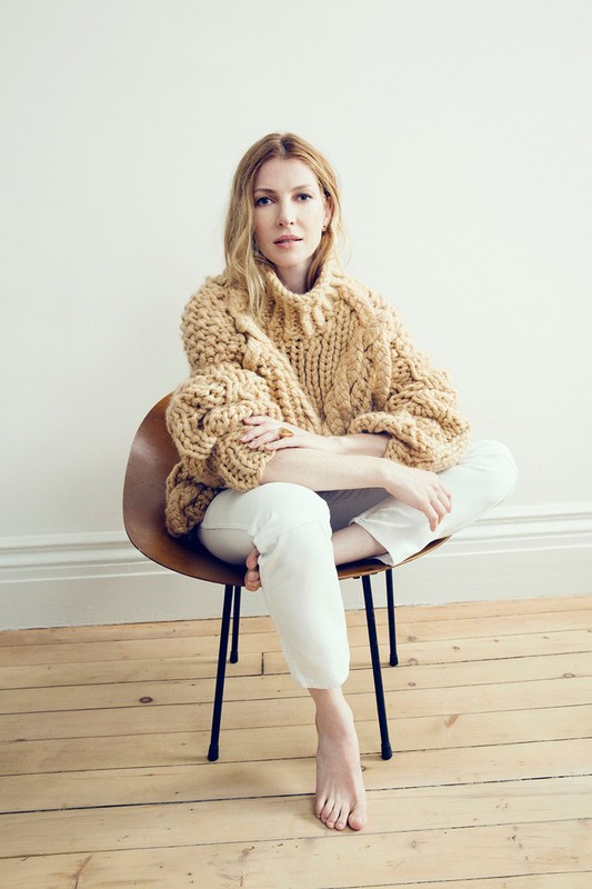 Portrait of woman in chunky knit sweater sitting on chair. Fair-skinned female with long blonde hair and bare feet in white studio.