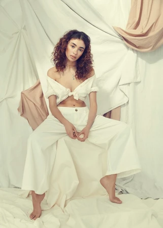A curly-haired woman in a white crop top and palazzo pants sits barefoot against draped fabrics in a fashion-forward studio shot.