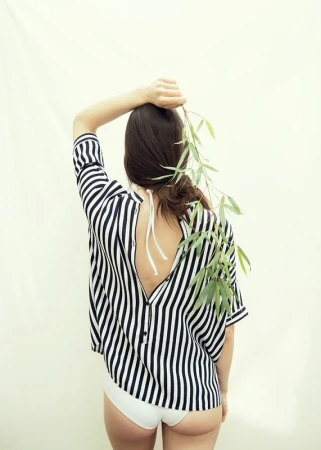 A woman in a striped blouse poses from the back, one arm raised, holding a branch with leaves against a plain background.