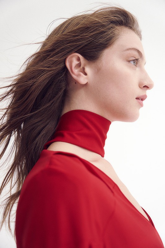 Woman with long brown hair poses in a red dress with matching high collar against a white background in a fashion headshot.