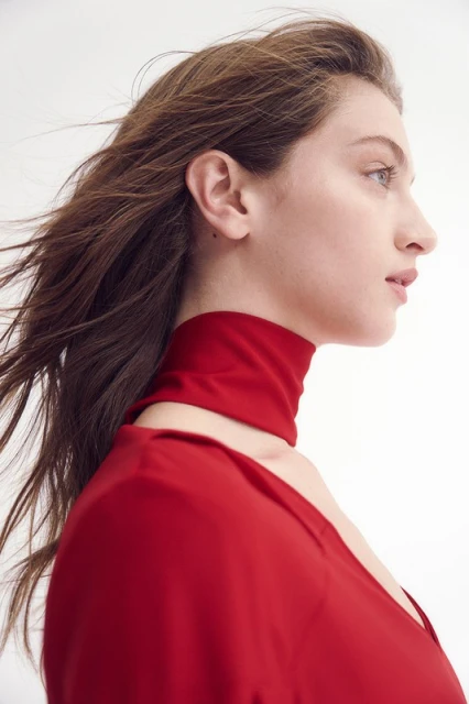 Woman with long brown hair poses in a red dress with matching high collar against a white background in a fashion headshot.