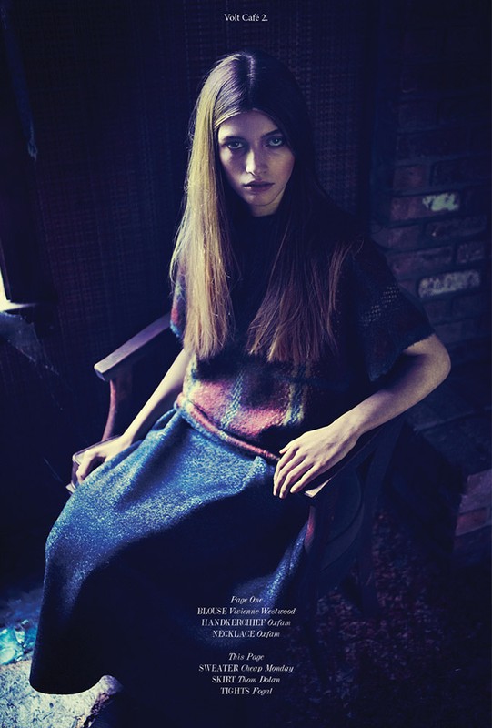 Fashion shot of a woman with long brown hair sitting on a chair, wearing a printed top and navy blue skirt indoors in dim lighting.