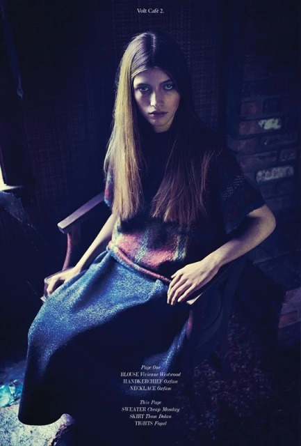 Fashion shot of a woman with long brown hair sitting on a chair, wearing a printed top and navy blue skirt indoors in dim lighting.