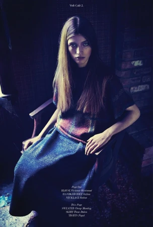 Fashion shot of a woman with long brown hair sitting on a chair, wearing a printed top and navy blue skirt indoors in dim lighting.