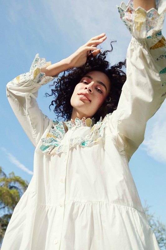 A woman in a white dress stands outside under a blue sky, looking up with her arms raised in a fashion style pose for an outdoor fashion shot.