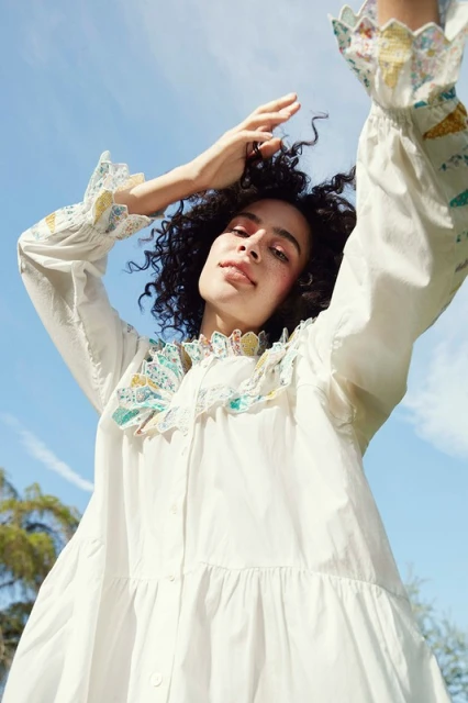 A woman in a white dress stands outside under a blue sky, looking up with her arms raised in a fashion style pose for an outdoor fashion shot.
