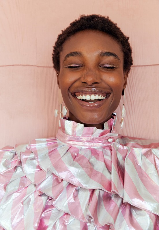 Close-up of an African-American woman with short hair laughing with closed eyes, wearing pink and silver ruffle top, earrings, against light pink background.