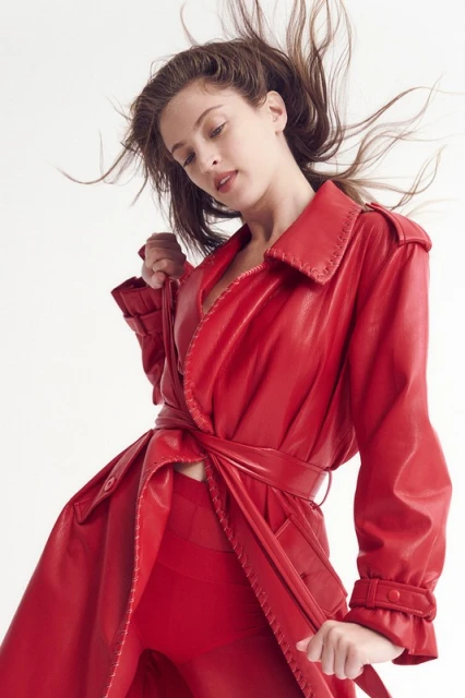 Young woman in motion poses in vibrant red leather trench coat and matching pants against a white background, fashion forward, studio lighting.