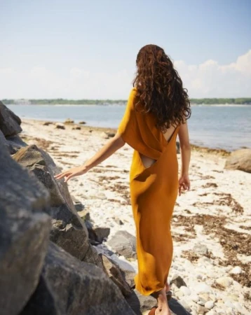 Woman with long curly brown hair wearing an amber yellow dress walking along a sandy beach near rocks.