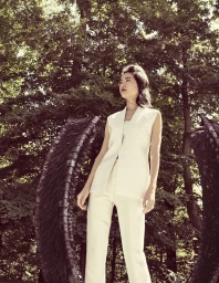 Woman wearing a white sleeveless vest and trousers standing outdoors near large arched sculptural structures with a forest background.