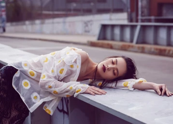 Woman in sheer white floral top with dark lips reclining on an outdoor concrete structure in Los Angeles.