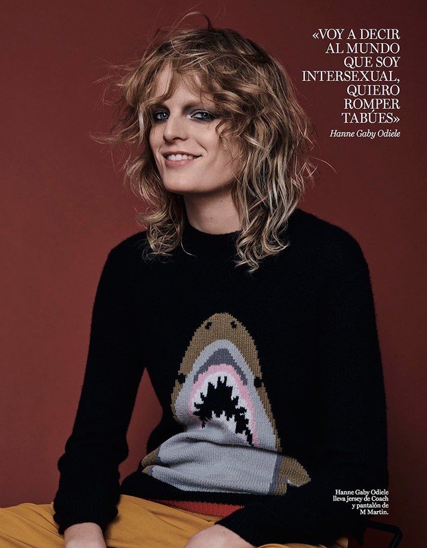 A woman with curly blonde hair smiles in a medium shot, wearing a black sweater with a shark design and ochre pants against a brown background.