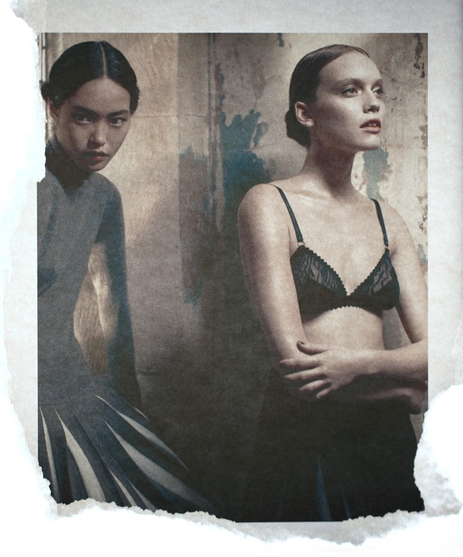Two women pose indoors against a textured wall, one in a grey dress, the other in black lingerie, vintage fashion, neutral color grading.