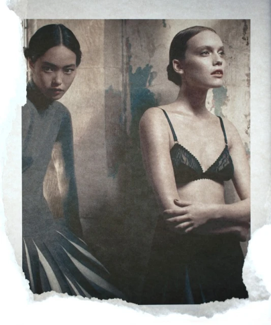 Two women pose indoors against a textured wall, one in a grey dress, the other in black lingerie, vintage fashion, neutral color grading.