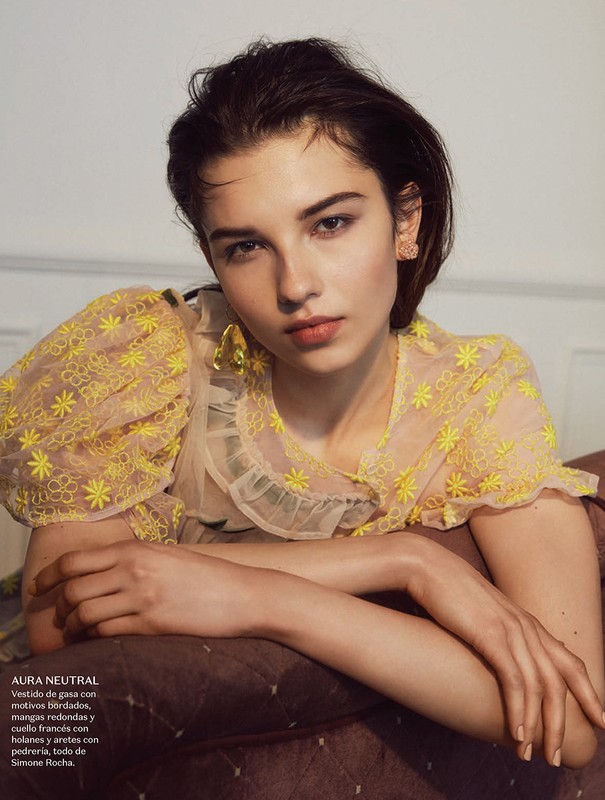 Brunette woman poses in a sheer yellow floral top indoors with a neutral expression, leaning on a brown quilted object.