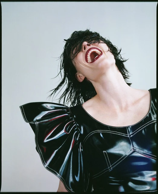 Woman with head tilted back and mouth open, wearing black vinyl dress with white stitching, photographed in a photo studio with soft lighting.