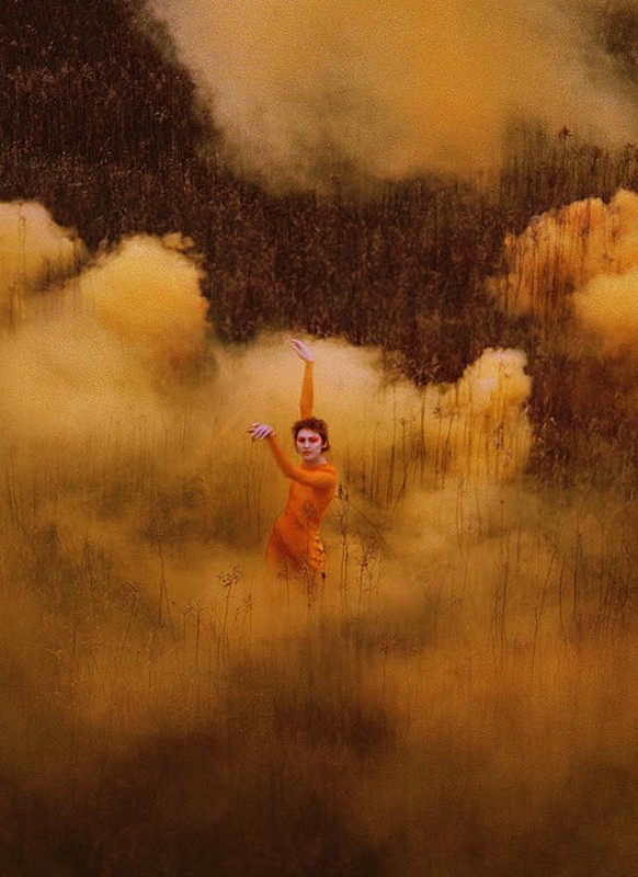 A person in an orange garment standing in a grassy field surrounded by a cloud of yellow and orange smoke or mist, facing the viewer.