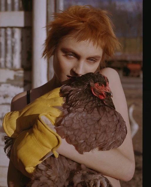 Portrait of a young woman with short orange hair holding a chicken in her arms while wearing yellow gloves in an outdoor setting.