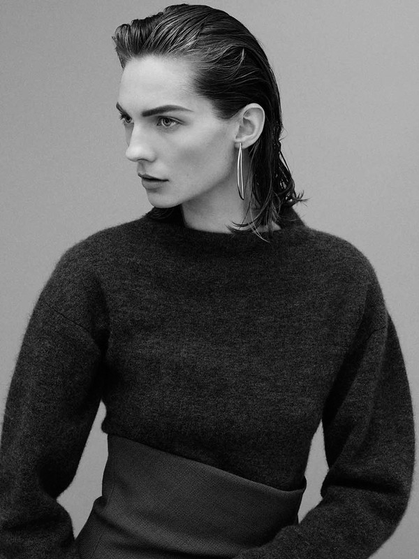 Monochrome portrait of a woman with slicked-back hair wearing a dark sweater and dark skirt, looking to the left, studio shot.