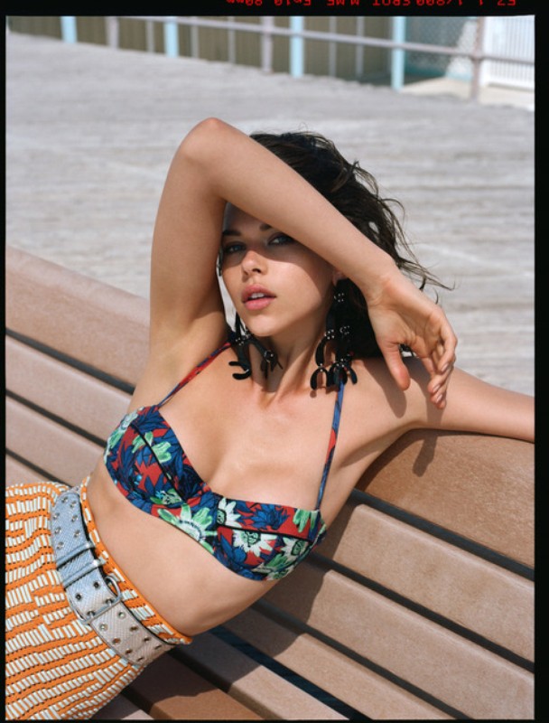 Brunette woman in floral bikini top reclines on boardwalk bench near beach in outdoor medium shot with a natural expression.