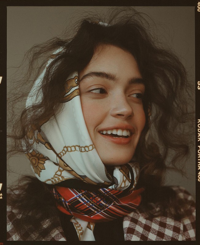 Smiling woman in a patterned headscarf and top looks off to the side in a close-up portrait taken in a photo studio.