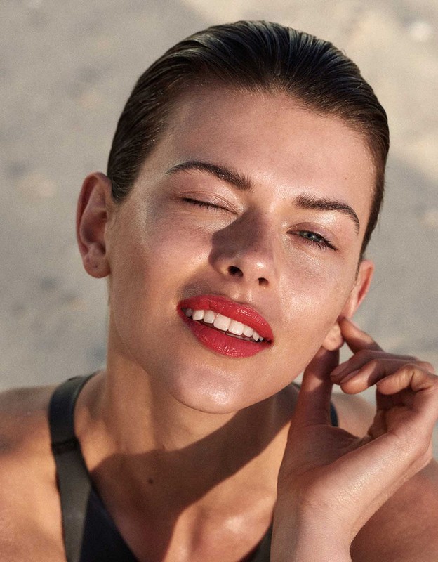 Close-up portrait of an adult female winking, with wet slicked-back hair, red lipstick, and wearing a black top against a blurred sandy background.