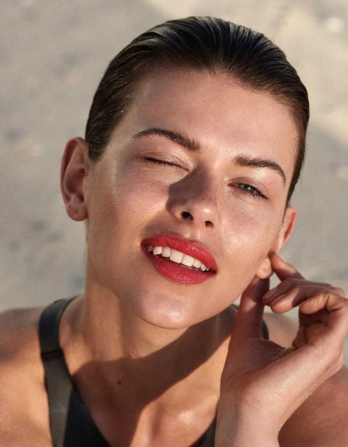 Close-up portrait of an adult female winking, with wet slicked-back hair, red lipstick, and wearing a black top against a blurred sandy background.