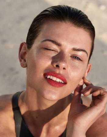 Close-up portrait of an adult female winking, with wet slicked-back hair, red lipstick, and wearing a black top against a blurred sandy background.