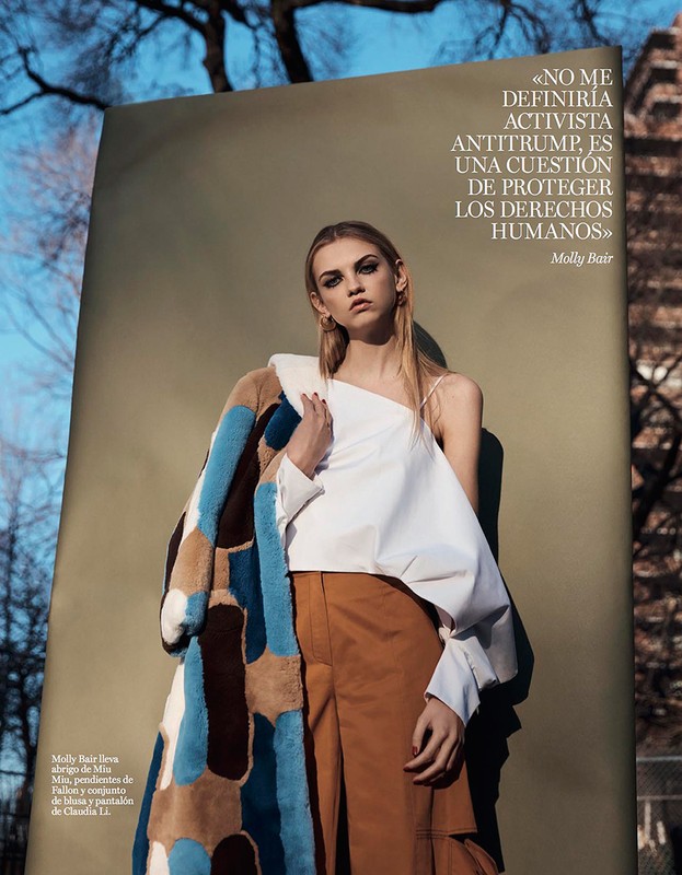 A woman in a white top and tan pants poses in front of a tan backdrop with an open coat. The image is a fashion editorial.