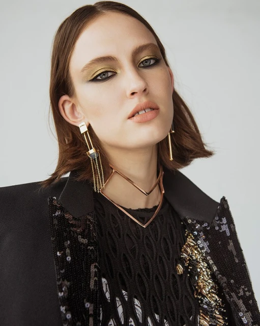Close-up of a young woman with olive eyeshadow and statement jewelry, wearing a sequined jacket, against a neutral backdrop.