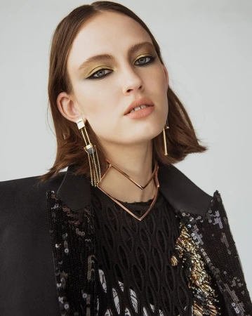 Close-up of a young woman with olive eyeshadow and statement jewelry, wearing a sequined jacket, against a neutral backdrop.
