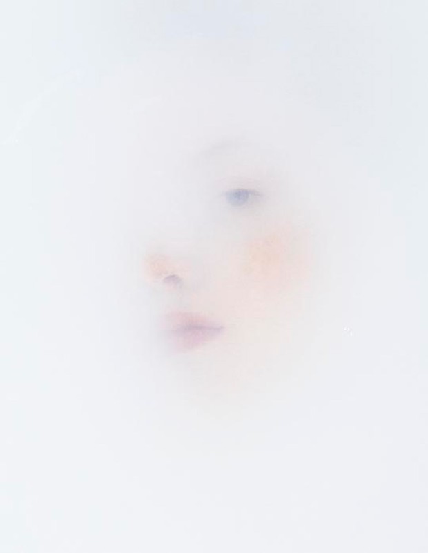 Close-up portrait of a fair-skinned adult female face, subtly visible through a diffused, soft-focus, ethereal white background.