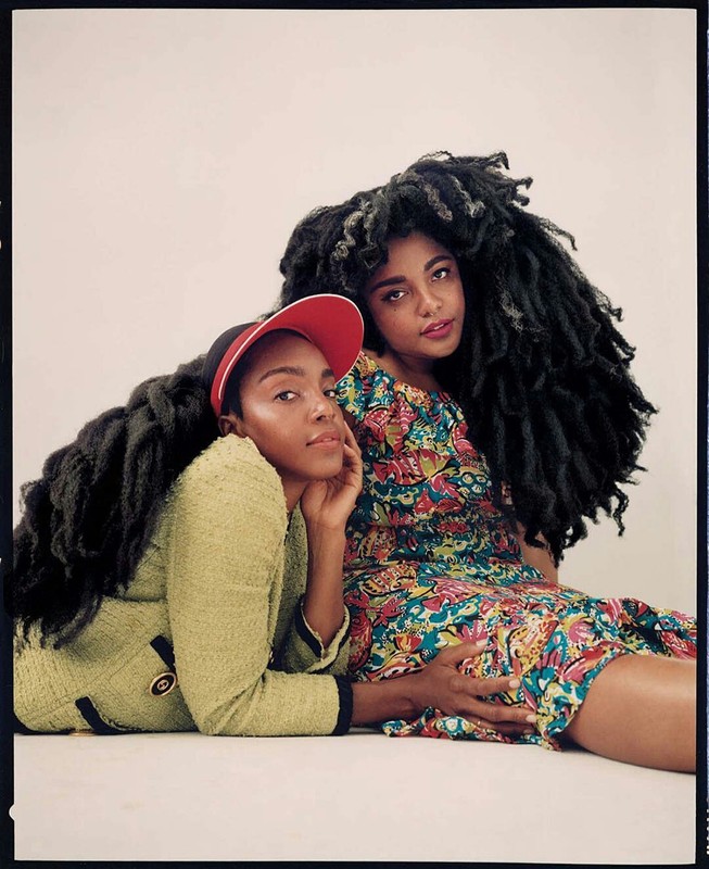 Two Black women with voluminous dark hair pose in a medium studio shot; one wears a red cap and chartreuse jacket, the other a floral dress.