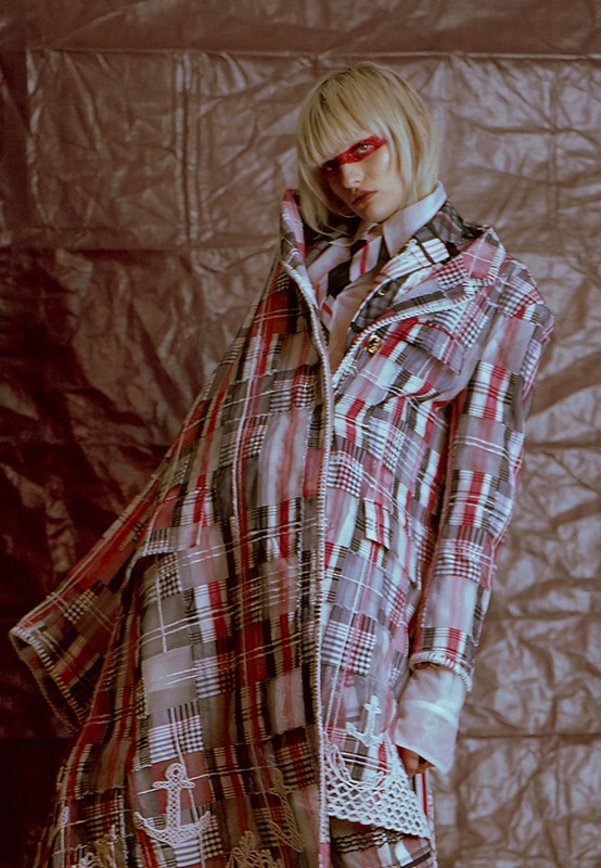 Fashion-forward female model in a stylish plaid coat with anchor details, posing against a textured metallic backdrop in a studio setting.