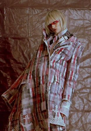 Fashion-forward female model in a stylish plaid coat with anchor details, posing against a textured metallic backdrop in a studio setting.