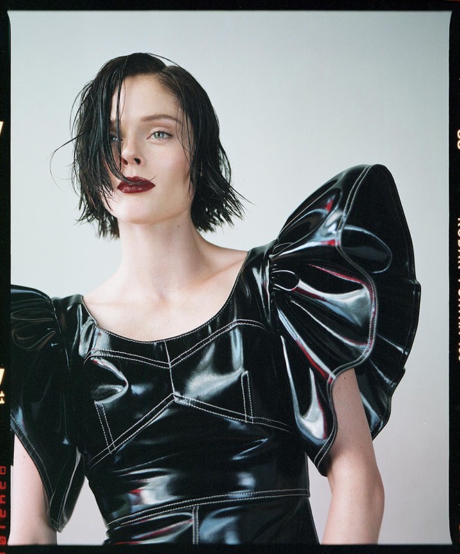 Fashion portrait of a woman with dark, wet hair and dark lipstick wearing a black, shiny dress with large, ruffled sleeves, studio shot.