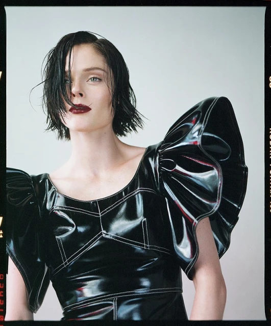 Fashion portrait of a woman with dark, wet hair and dark lipstick wearing a black, shiny dress with large, ruffled sleeves, studio shot.