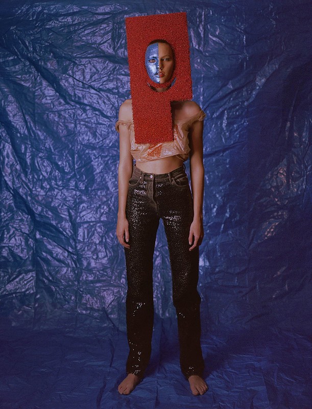Fashion portrait of a barefoot adult woman with blue face paint, wearing a red rectangular headpiece and sequined jeans in a photo studio.