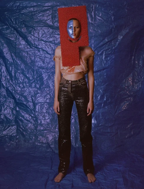 Fashion portrait of a barefoot adult woman with blue face paint, wearing a red rectangular headpiece and sequined jeans in a photo studio.