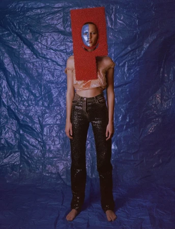 Fashion portrait of a barefoot adult woman with blue face paint, wearing a red rectangular headpiece and sequined jeans in a photo studio.