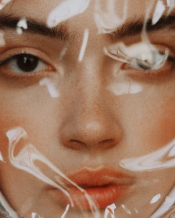 Close-up portrait of a young adult female with light skin, brown eyes, and freckles, partially obscured by transparent plastic wrap.