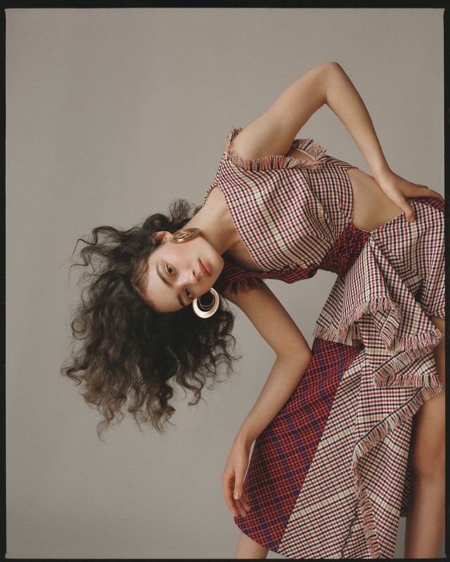 Portrait of an adult female fashion model with curly hair in a plaid dress posing in a studio with a neutral grey background.