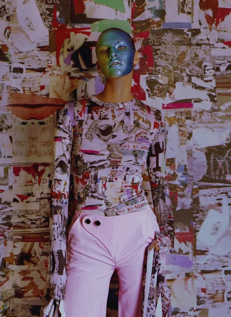 Fashionable adult model with iridescent makeup poses in front of a collage-style wall wearing a long-sleeved shirt and light pink pants.