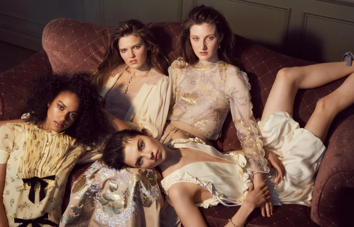 Four female models in patterned dresses lounging together on a plush mauve button-tufted velvet sofa in an interior setting.