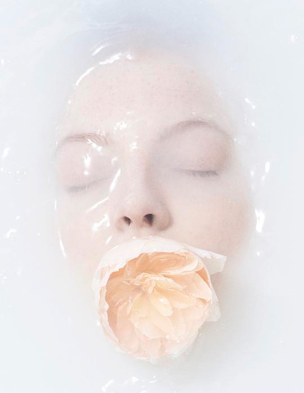 Woman submerged in water with eyes closed, holding a light orange rose in her mouth, creating a serene and ethereal beauty portrait.